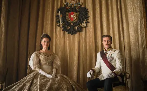 La principessa Sissi e l'imperatore Francesco Giuseppe nella serie tv, seduti su poltrone dorate, con uno stemma imperiale sullo sfondo.