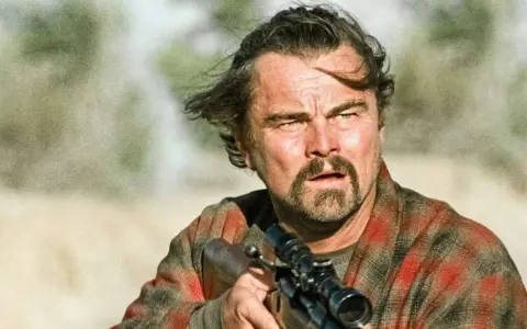 Leonardo DiCaprio con fucile, in un film di sopravvivenza. Capelli mossi dal vento, sguardo intenso, camicia a quadri.