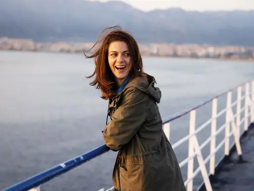 Una donna ride felice sul ponte di una nave, con la costa sullo sfondo. Sembra una scena da un **dieci minuti film** di avventura.