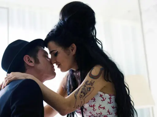 Blake Fielder-Civil, marito di Amy Winehouse, la bacia teneramente. Lei ha i capelli neri raccolti e indossa un top con ancore.