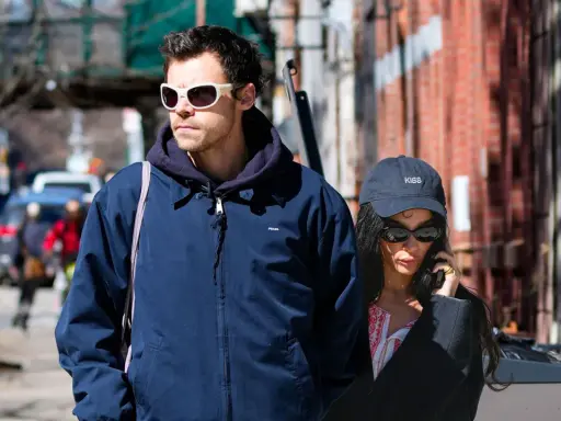 Harry Styles e la sua fidanzata passeggiano per strada, lui con occhiali bianchi, lei al telefono con un cappellino "KISS".