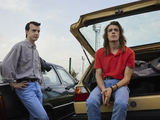 Due ragazzi in jeans e camicie, appoggiati a due auto d'epoca. Scena dalla serie tv 883.
