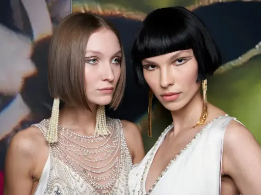 Due modelle con un sbarazzino caschetto corto sfilato, una con capelli castani e l'altra con capelli neri, sfoggiano orecchini pendenti e abiti decorati.
