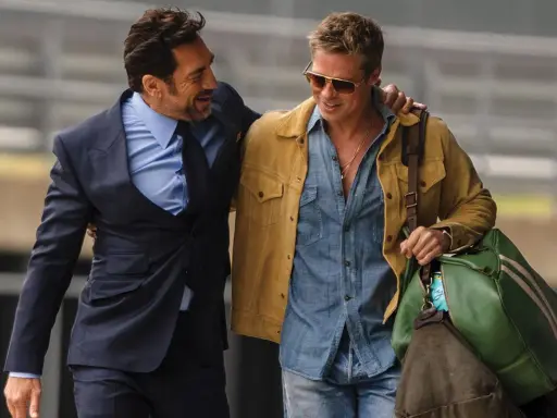 Brad Pitt 2024, con giacca gialla e camicia di jeans, cammina con un amico in completo scuro.