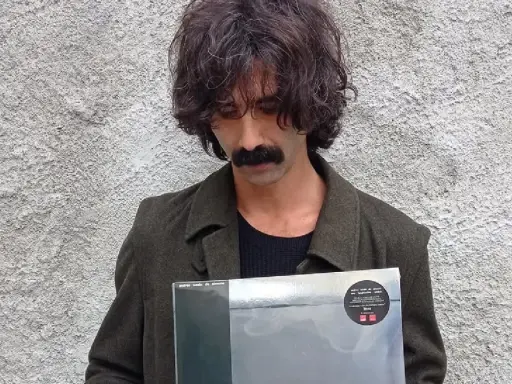 Andrea Laszlo De Simone con un disco in mano, con il suo iconico baffo e capelli ricci, davanti a un muro grezzo.