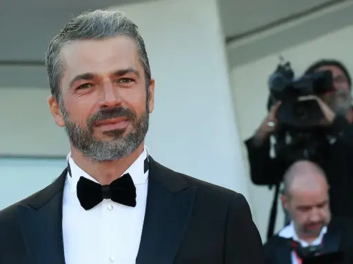 Uomo con capelli bianchi e barba, indossa un abito elegante con papillon nero. Dietro di lui, un operatore riprende con una telecamera, quasi a voler dimostrare che i capelli bianchi possono tornare neri.