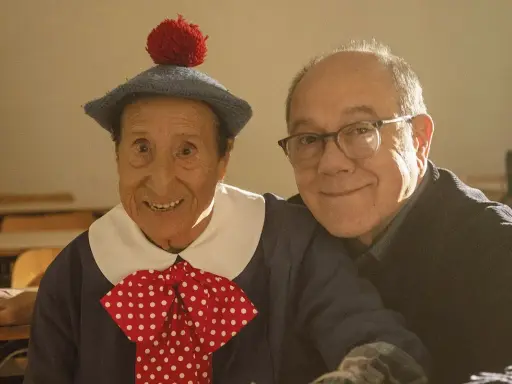 Un uomo con un cappello blu e un fiocco a pois rossi, sorride accanto a un uomo con gli occhiali. Un momento di gioia nella vita da Carlo.