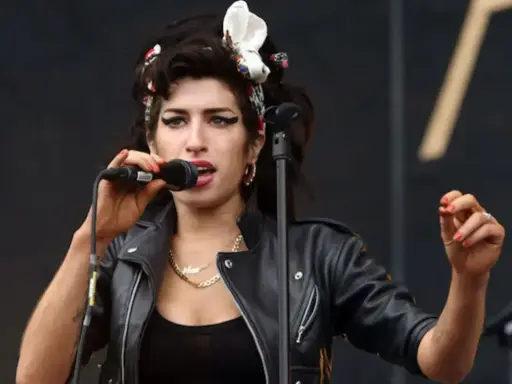 Amy Winehouse magra, con la sua iconica acconciatura e trucco, canta al microfono con passione. Indossa una giacca di pelle nera.