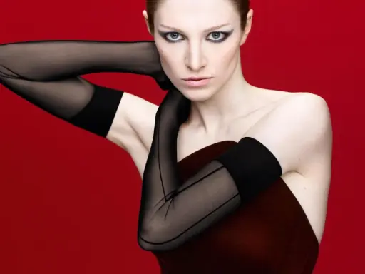Hunter Schafer, prima donna con guanti neri velati e trucco drammatico, posa con le braccia incrociate su sfondo rosso.