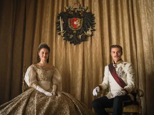 La principessa Sissi e l'imperatore Francesco Giuseppe nella serie tv, seduti su poltrone dorate, con uno stemma imperiale sullo sfondo.