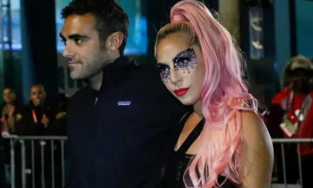 Lady Gaga con il suo fidanzato, Christian Carino, durante un evento. Lei indossa un trucco scintillante e capelli rosa.