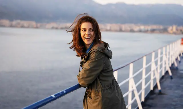 Una donna ride felice sul ponte di una nave, con la costa sullo sfondo. Sembra una scena da un **dieci minuti film** di avventura.