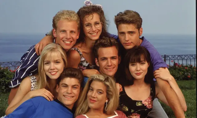 Il cast di Beverly Hills 90210, un gruppo di amici sorridenti con lo sfondo del mare.