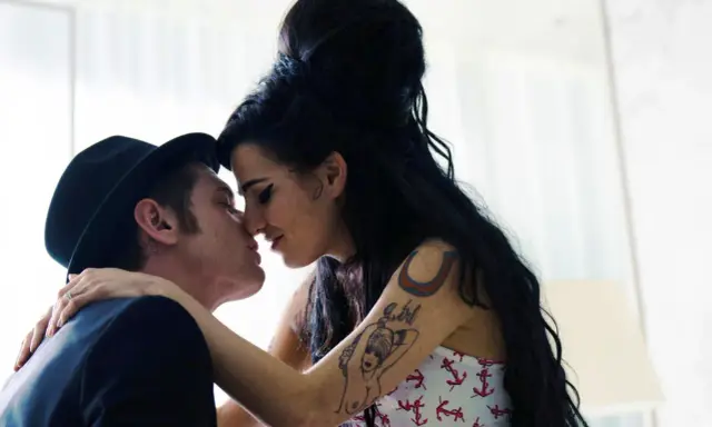 Blake Fielder-Civil, marito di Amy Winehouse, la bacia teneramente. Lei ha i capelli neri raccolti e indossa un top con ancore.