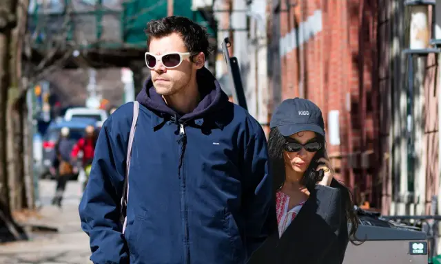 Harry Styles e la sua fidanzata passeggiano per strada, lui con occhiali bianchi, lei al telefono con un cappellino "KISS".