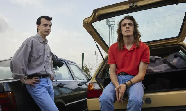 Due ragazzi in jeans e camicie, appoggiati a due auto d'epoca. Scena dalla serie tv 883.