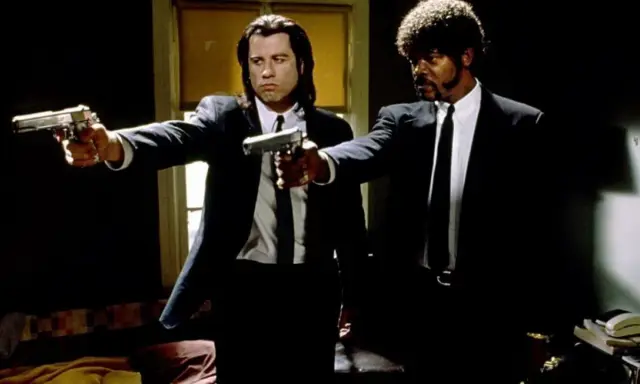 Due uomini in abito scuro puntano pistole, iconica scena da un film anni 90.