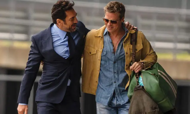 Brad Pitt 2024, con giacca gialla e camicia di jeans, cammina con un amico in completo scuro.