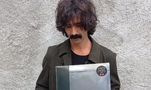 Andrea Laszlo De Simone con un disco in mano, con il suo iconico baffo e capelli ricci, davanti a un muro grezzo.