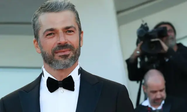 Uomo con capelli bianchi e barba, indossa un abito elegante con papillon nero. Dietro di lui, un operatore riprende con una telecamera, quasi a voler dimostrare che i capelli bianchi possono tornare neri.