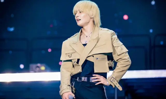 Uomo con capelli biondi e taglio wolf cut, indossa una giacca beige oversize e pantaloni neri.