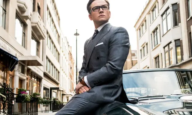 Taron Egerton, parte del cast di Kingsman 2, posa con eleganza su un'auto d'epoca, vestito in un impeccabile abito grigio.