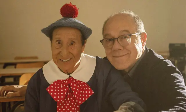 Un uomo con un cappello blu e un fiocco a pois rossi, sorride accanto a un uomo con gli occhiali. Un momento di gioia nella vita da Carlo.