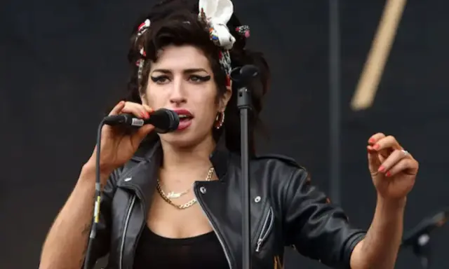 Amy Winehouse magra, con la sua iconica acconciatura e trucco, canta al microfono con passione. Indossa una giacca di pelle nera.
