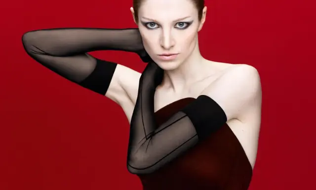 Hunter Schafer, prima donna con guanti neri velati e trucco drammatico, posa con le braccia incrociate su sfondo rosso.