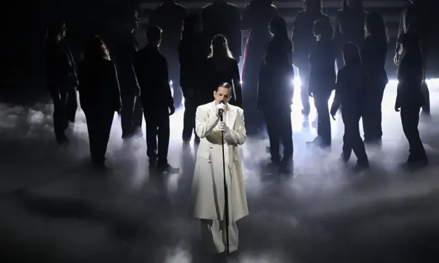 Achille Lauro, vestito di bianco, canta sul palco circondato da un coro in controluce, un'immagine che evoca un sentimento di essere perdutamente innamorato.