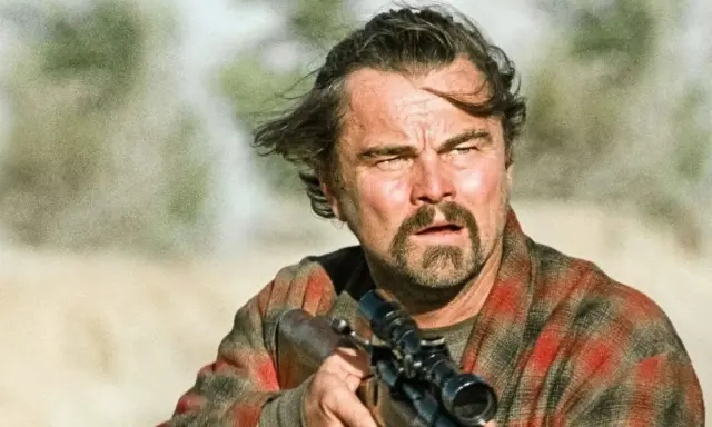 Leonardo DiCaprio con fucile, in un film di sopravvivenza. Capelli mossi dal vento, sguardo intenso, camicia a quadri.
