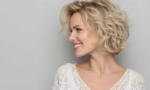 Donna sorridente con capelli biondi mossi, taglio **dietro sfumato caschetto corto**, indossa un top bianco in pizzo.