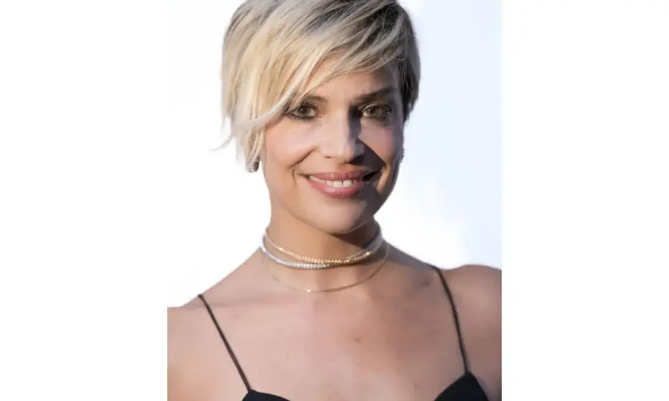 Donna con capelli cortissimi biondi, look 2026, indossa un abito nero e collane scintillanti.