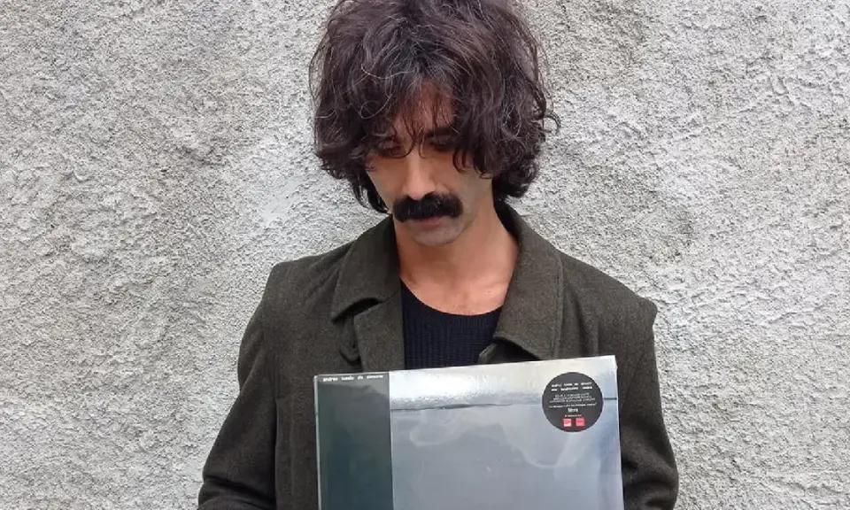 Andrea Laszlo De Simone con un disco in mano, con il suo iconico baffo e capelli ricci, davanti a un muro grezzo.