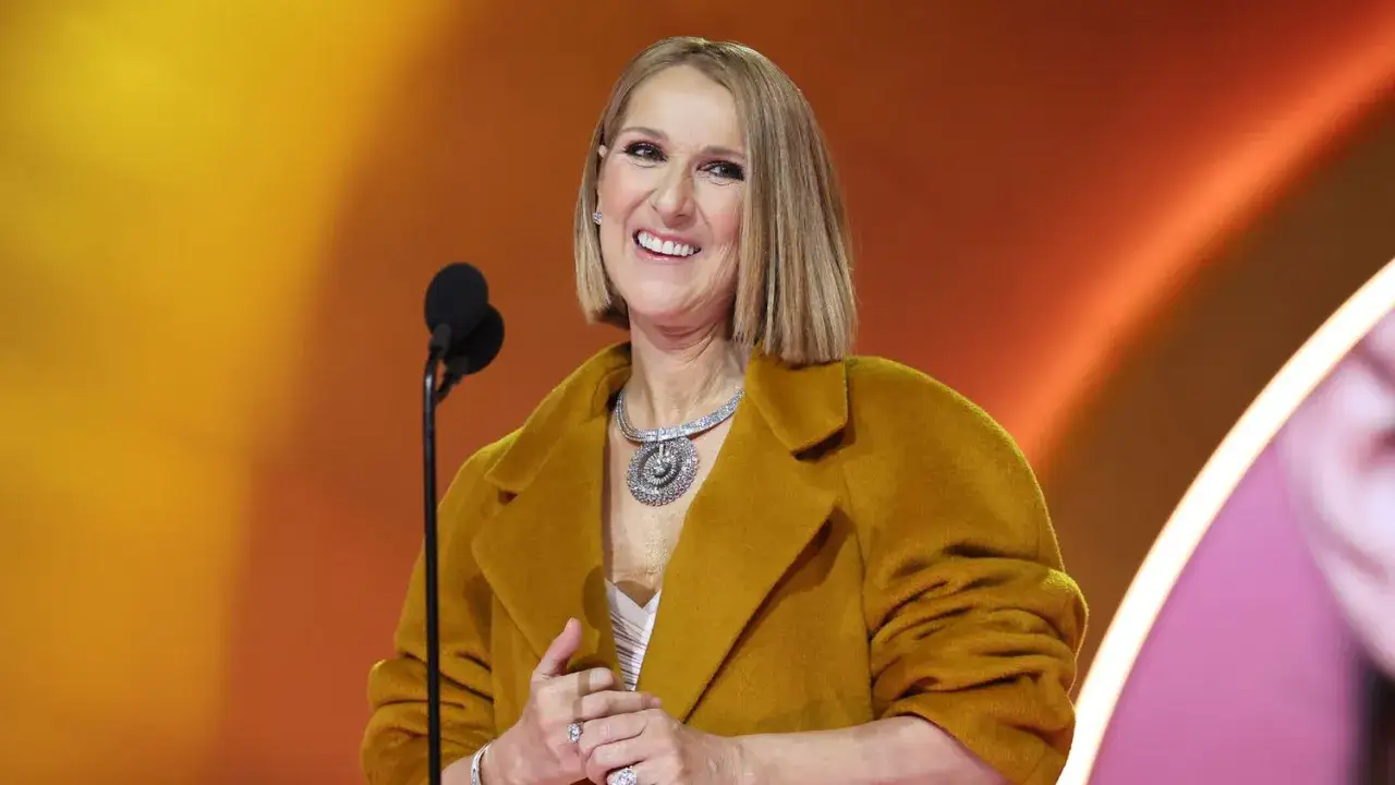 Celine Dion sorride con un cappotto ocra e un collier scintillante, mentre affronta la sua malattia con coraggio.