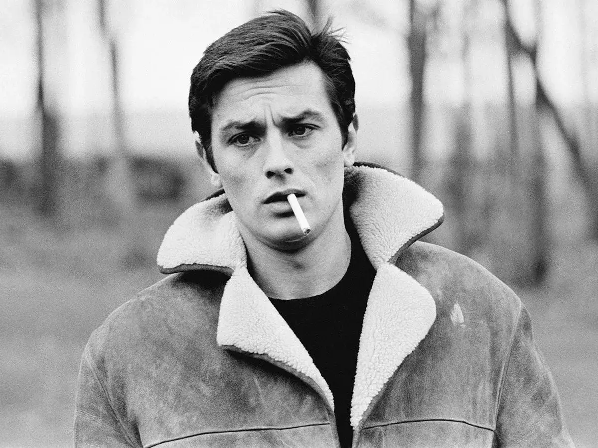 Alain Delon, iconico fisico, fuma una sigaretta con sguardo intenso, indossa una giacca di montone con colletto di pelliccia.