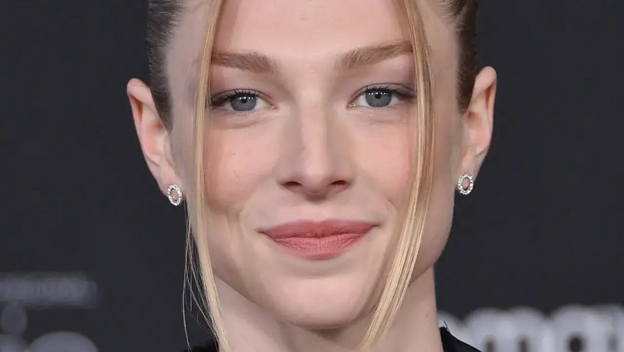Primo piano di Hunter Schafer, con occhi azzurri penetranti e un sorriso accennato. Indossa orecchini a cerchio scintillanti.