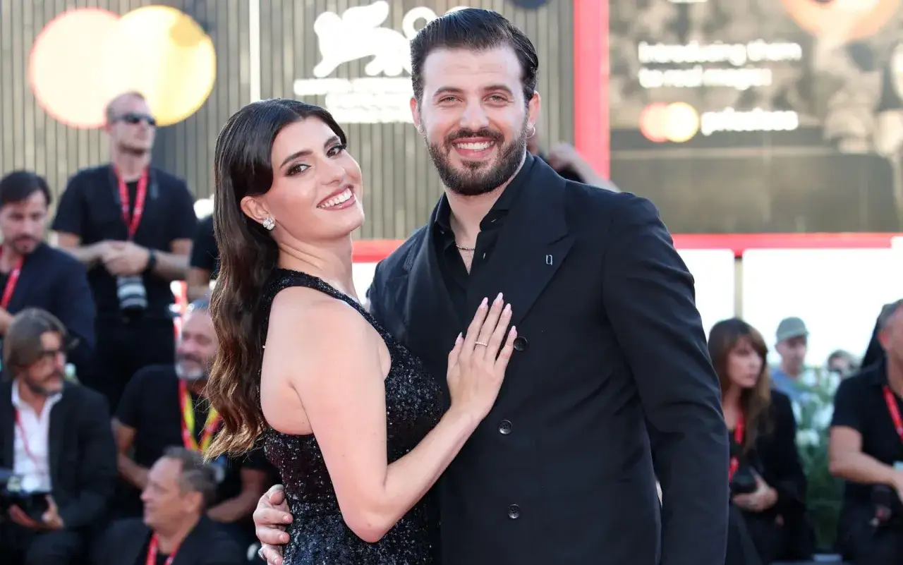 Gaetano Migliaccio e la sua compagna sorridono sul red carpet. Lei indossa un abito scintillante, lui un completo scuro.