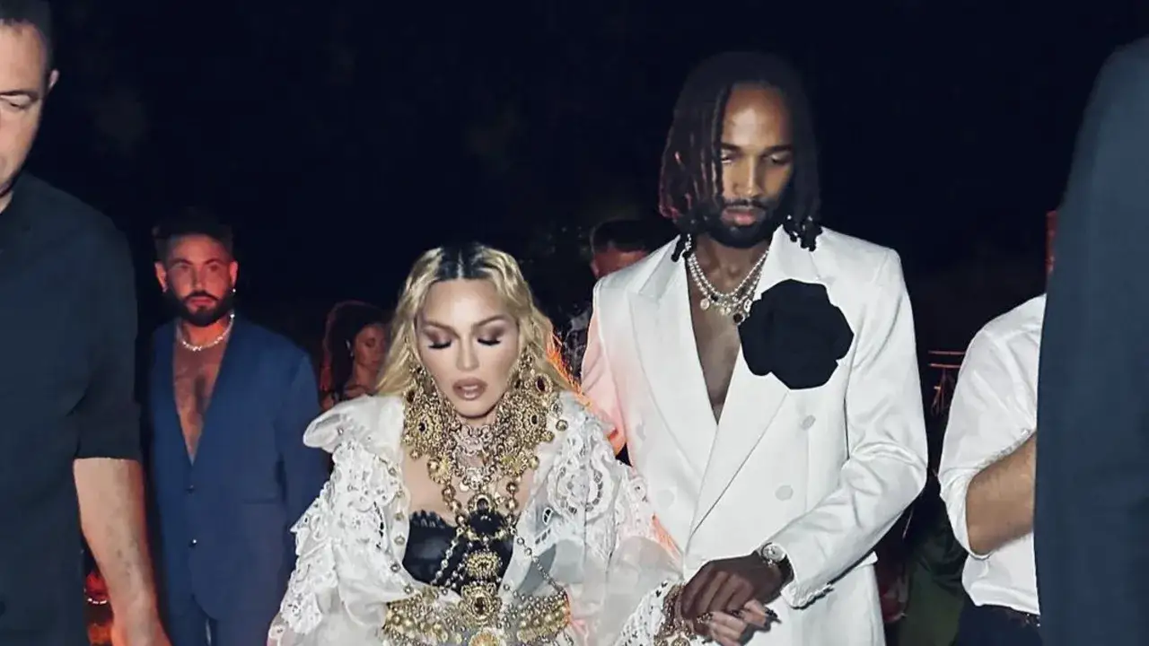 Madonna, con un sontuoso abito bianco e gioielli dorati, cammina mano nella mano con il suo compagno, vestito di bianco con una rosa nera.