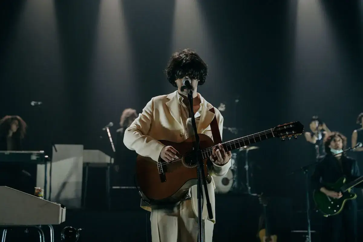 Andrea Laszlo De Simone canta e suona la chitarra su un palco illuminato, circondato dalla sua band.