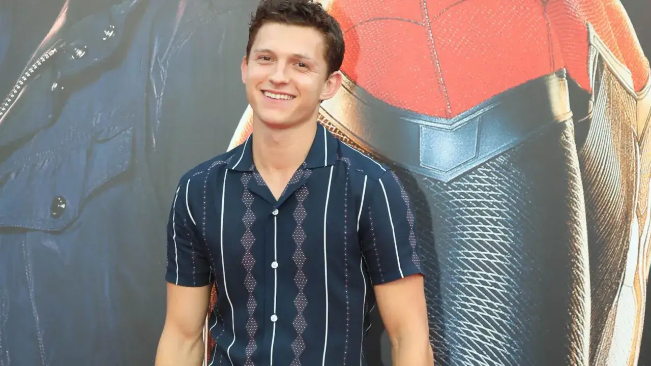 Tom Holland sorride, con uno sfondo che ricorda il costume di Spider-Man. Chissà quanti anni ha Tom Holland oggi?