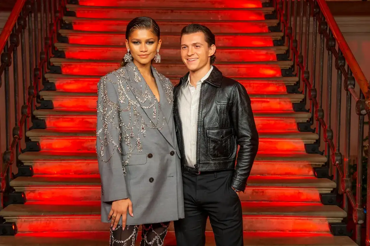 Tom Holland e Zendaya sorridono su una scala illuminata di rosso. Zendaya indossa un blazer grigio decorato, Tom un giubbotto di pelle.