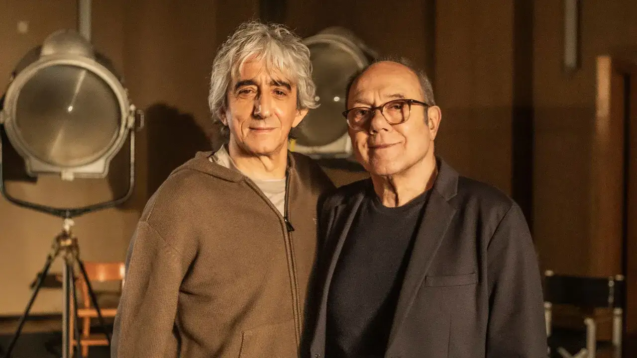 Due uomini sorridono, uno con capelli grigi e felpa marrone, l'altro con occhiali e giacca scura. Sembra una scena di vita da Carlo, forse sul set.