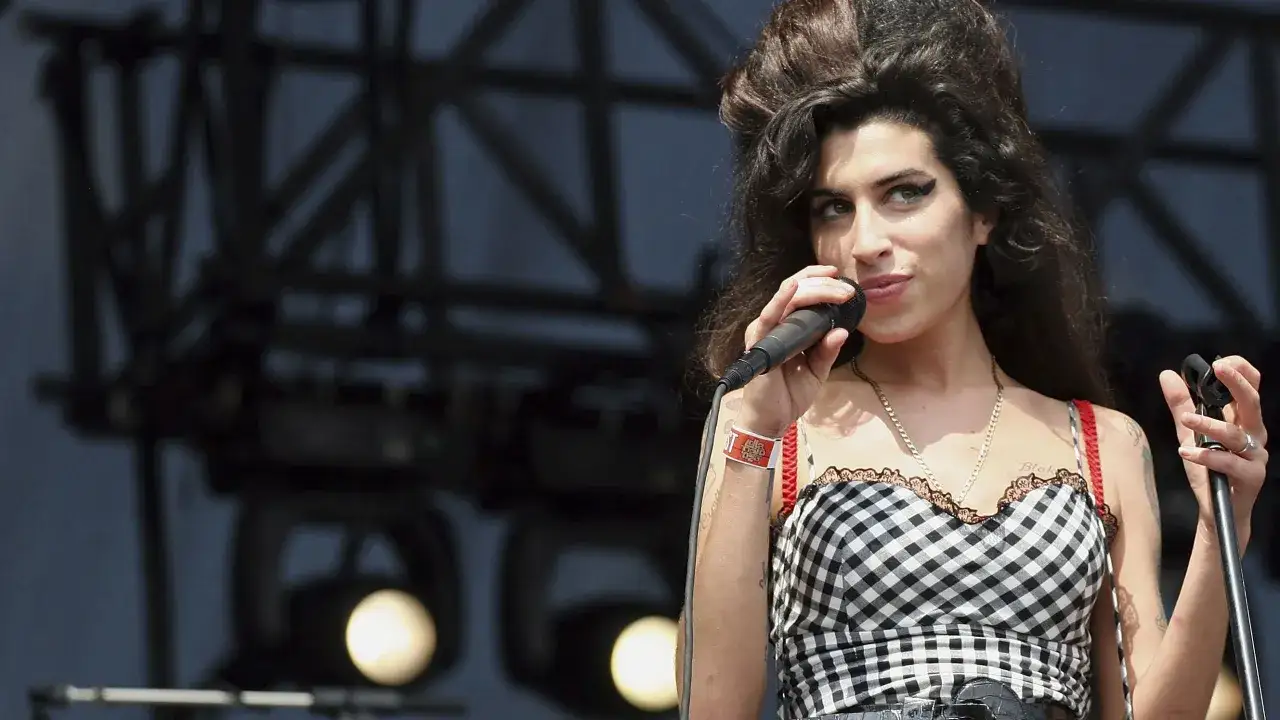 Amy Winehouse magra, con il suo iconico eyeliner, canta al microfono con un vestito a quadri.