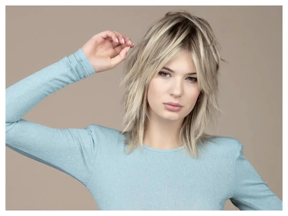 Modella con un audace taglio scalato medio corto, capelli biondi mossi e un top azzurro scintillante.