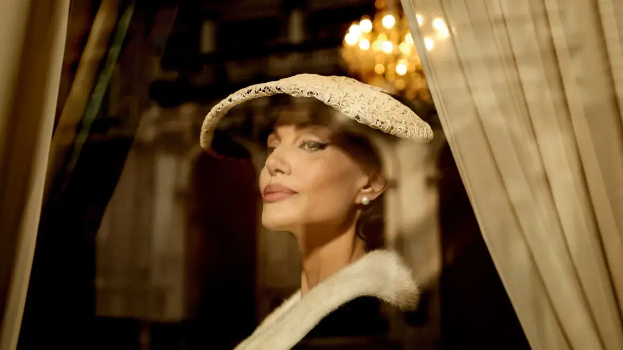 Angelina Jolie nei panni di Maria Callas, con un cappello di paglia e una pelliccia, riflessa in una vetrina.