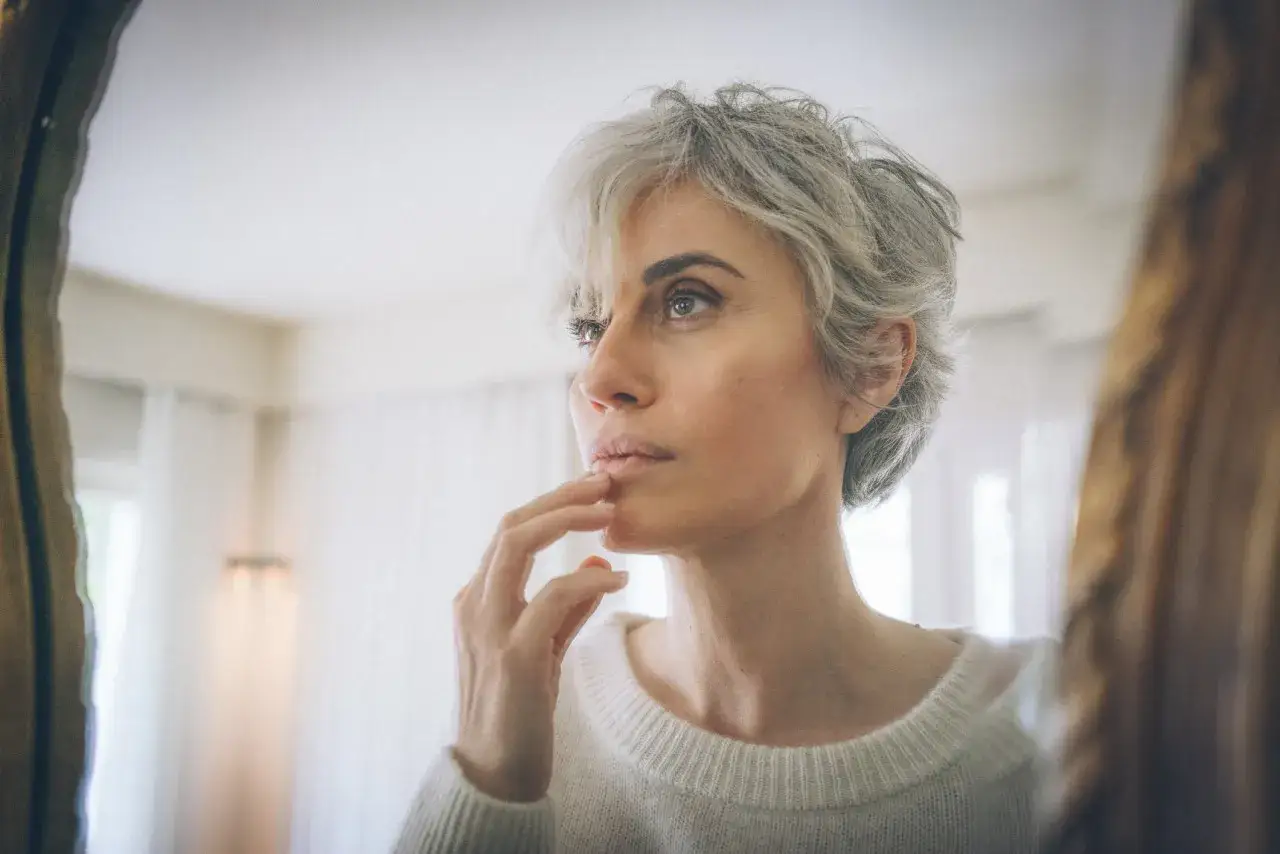 Donna con capelli bianchi guarda nello specchio, pensierosa. Chissà se i capelli bianchi possono tornare neri, un sogno di giovinezza.