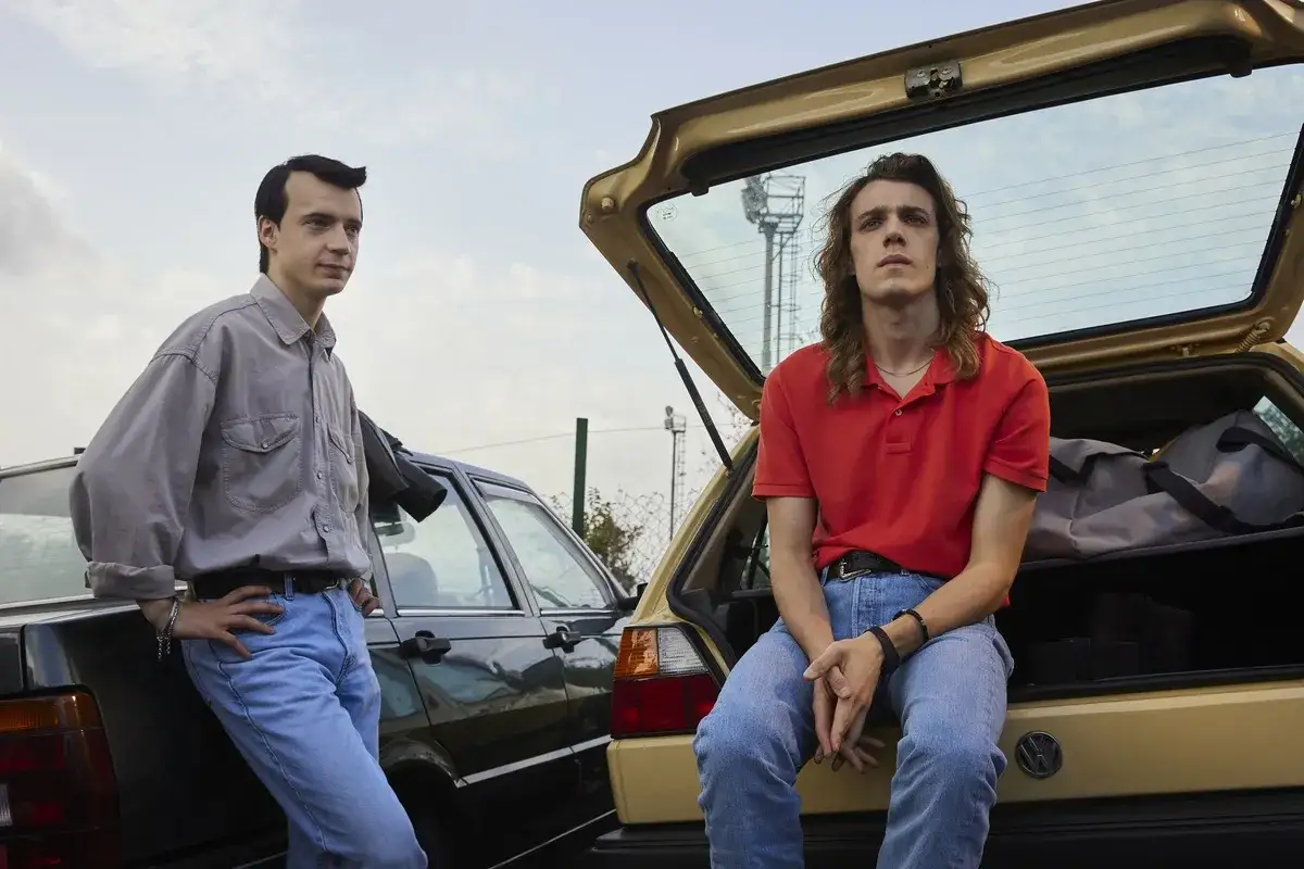 Due ragazzi, uno appoggiato a un'auto scura, l'altro seduto nel bagagliaio aperto di un'altra, sembrano pronti per un viaggio. Scena dalla serie tv 883.