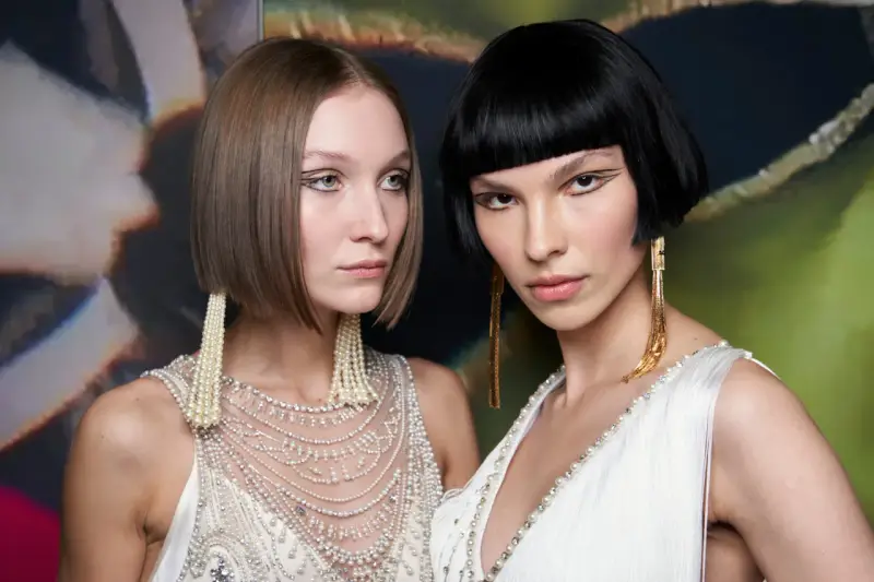 Due modelle con un sbarazzino caschetto corto sfilato, una con capelli castani e l'altra con capelli neri, sfoggiano orecchini pendenti e abiti decorati.